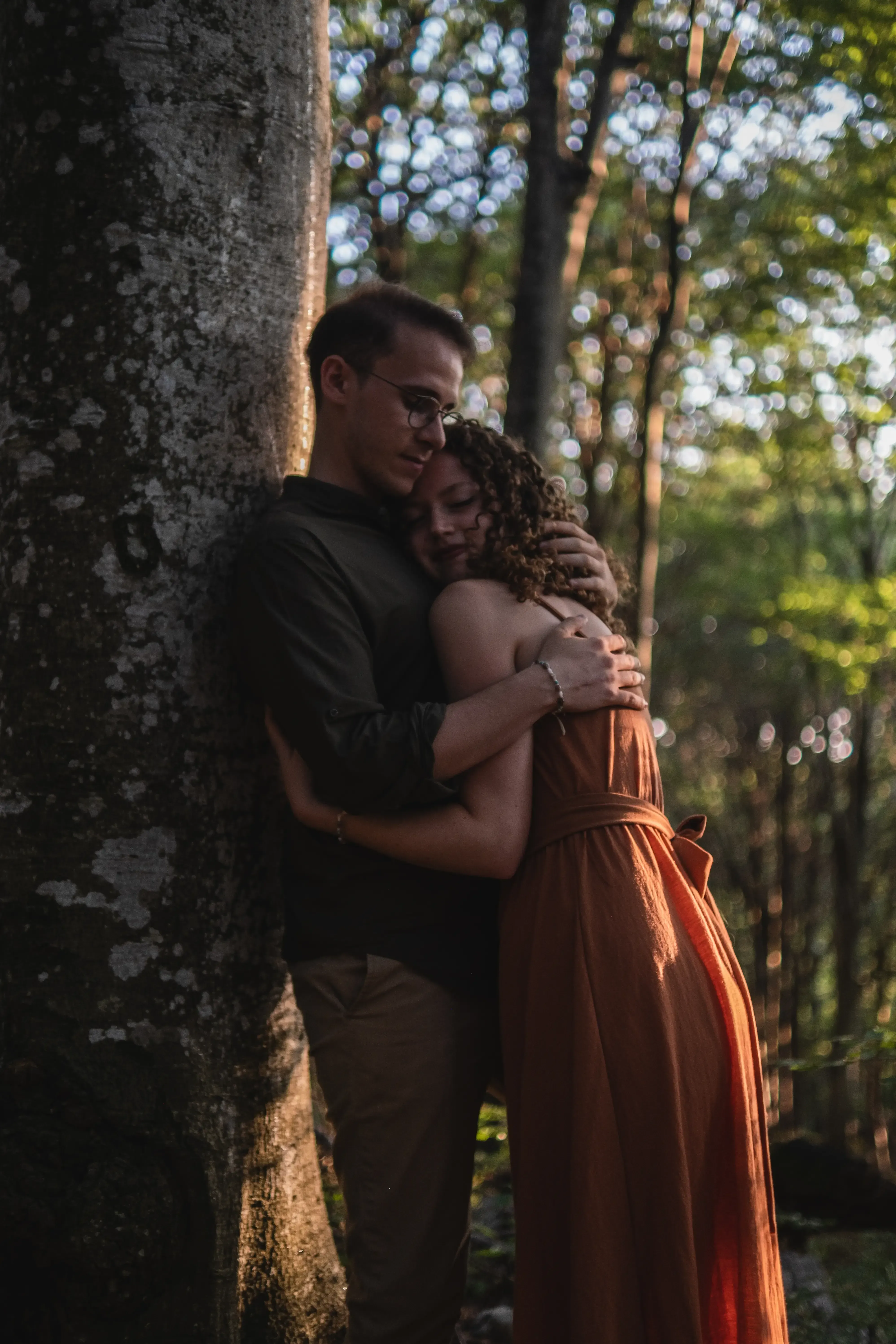 Nicolò leaning against a tree while hugging Greta in the forest.