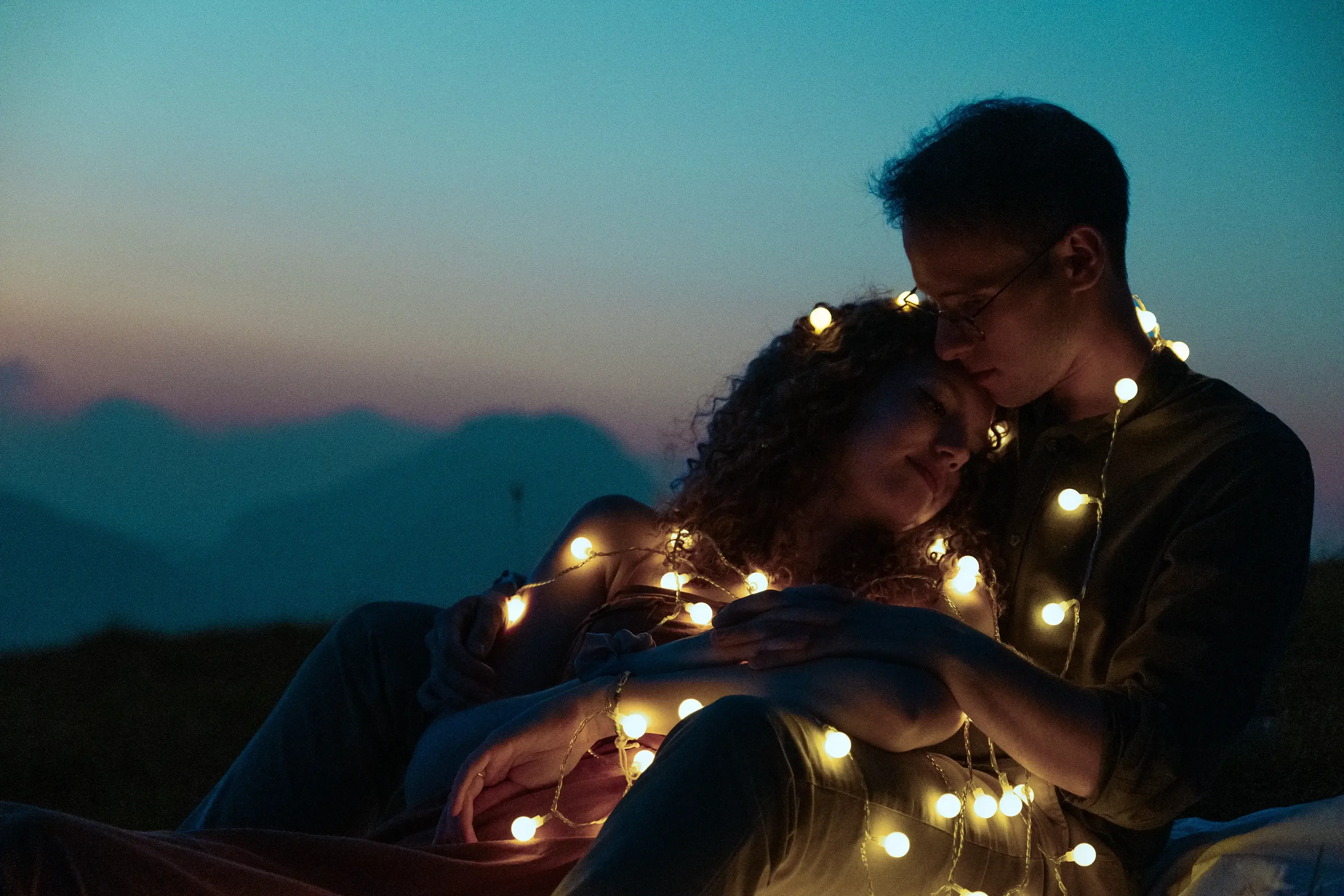 Nicolò and Greta hugging at night, lit by warm, delicate string lights in the mountain meadow.