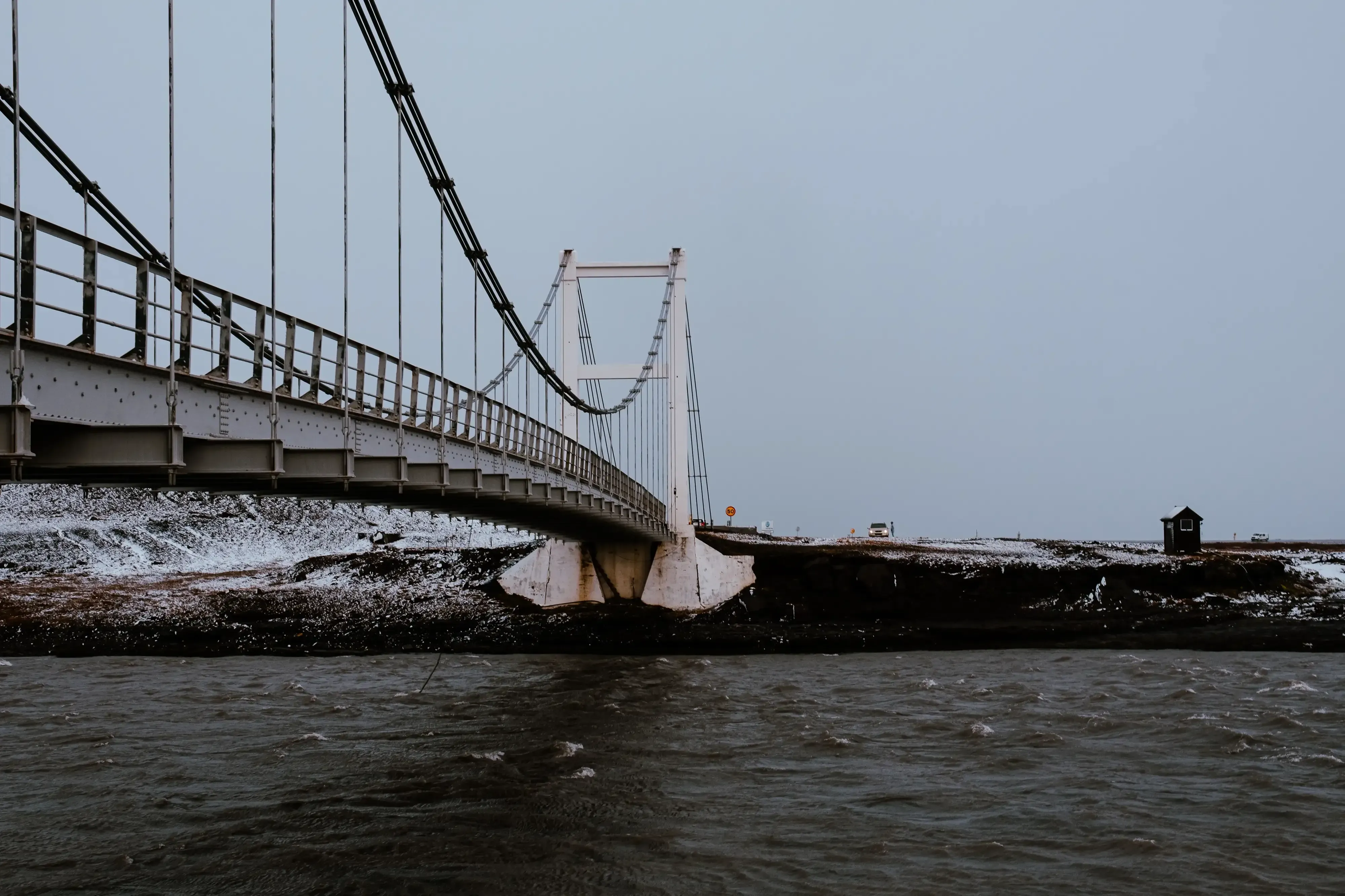 A clean, white arched or truss bridge spanning a river in the Icelandic landscape, viewed from the side.