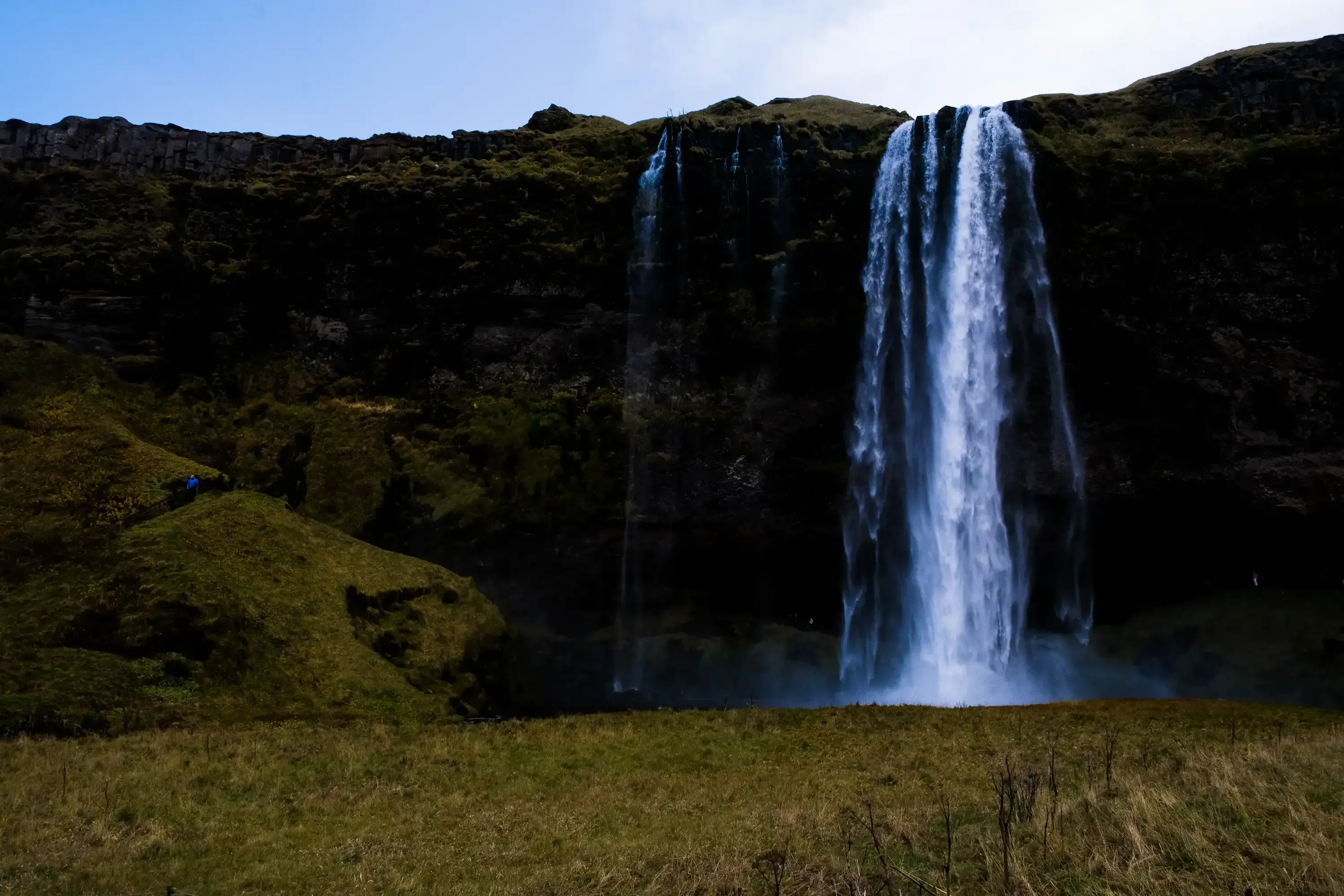 A wide, distant view capturing the entire height and surrounding landscape of the Seljalandsfoss waterfall.