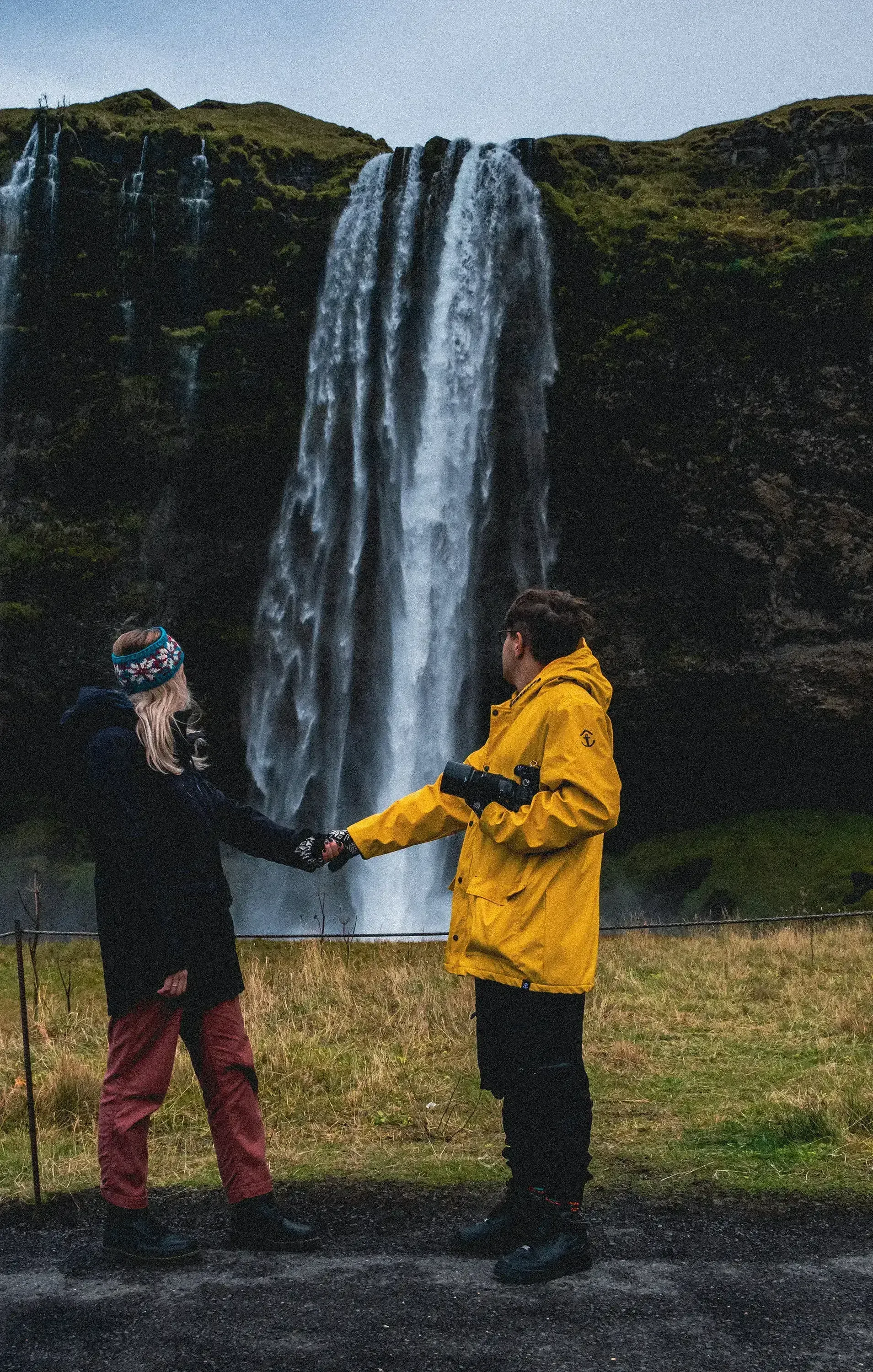 Ieser and Letizia standing together in front of the Seljalandsfoss waterfall, possibly near the entrance to the path behind it.