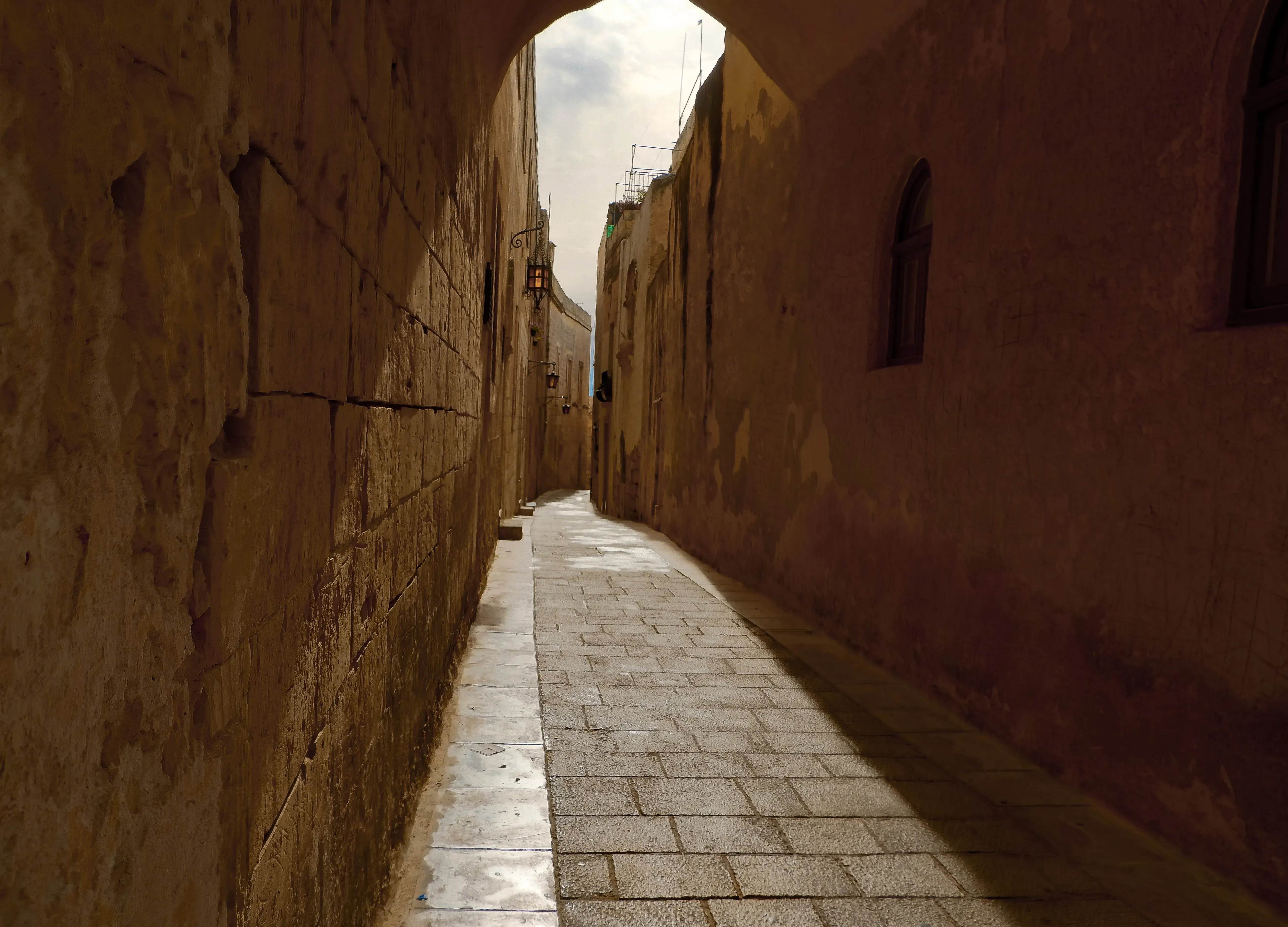 Narrow, golden-hued street in the fortified city of Mdina illuminated by sunlight.