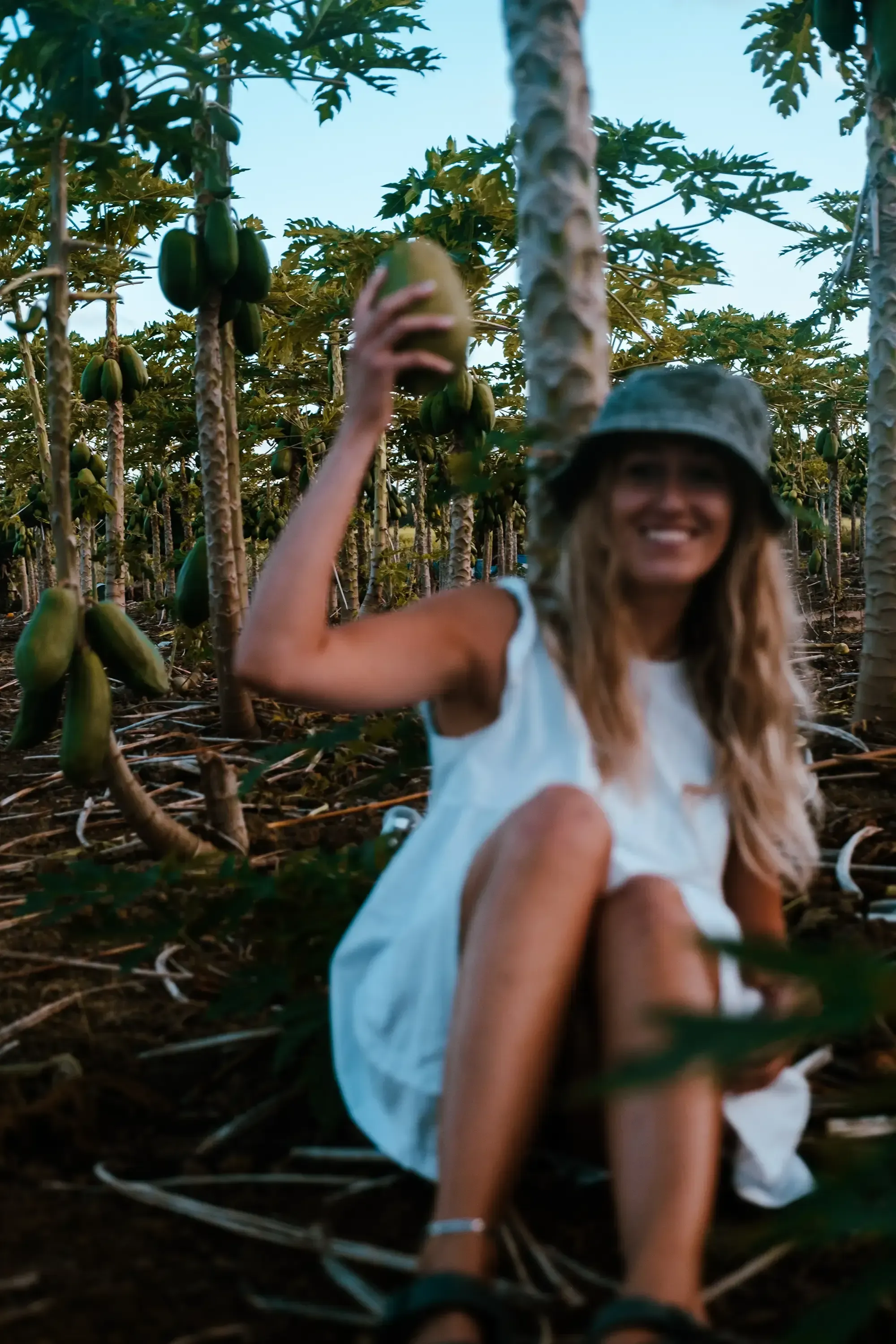 Letizia holding a papaya while sitting amidst a field of various tropical plants or crops.