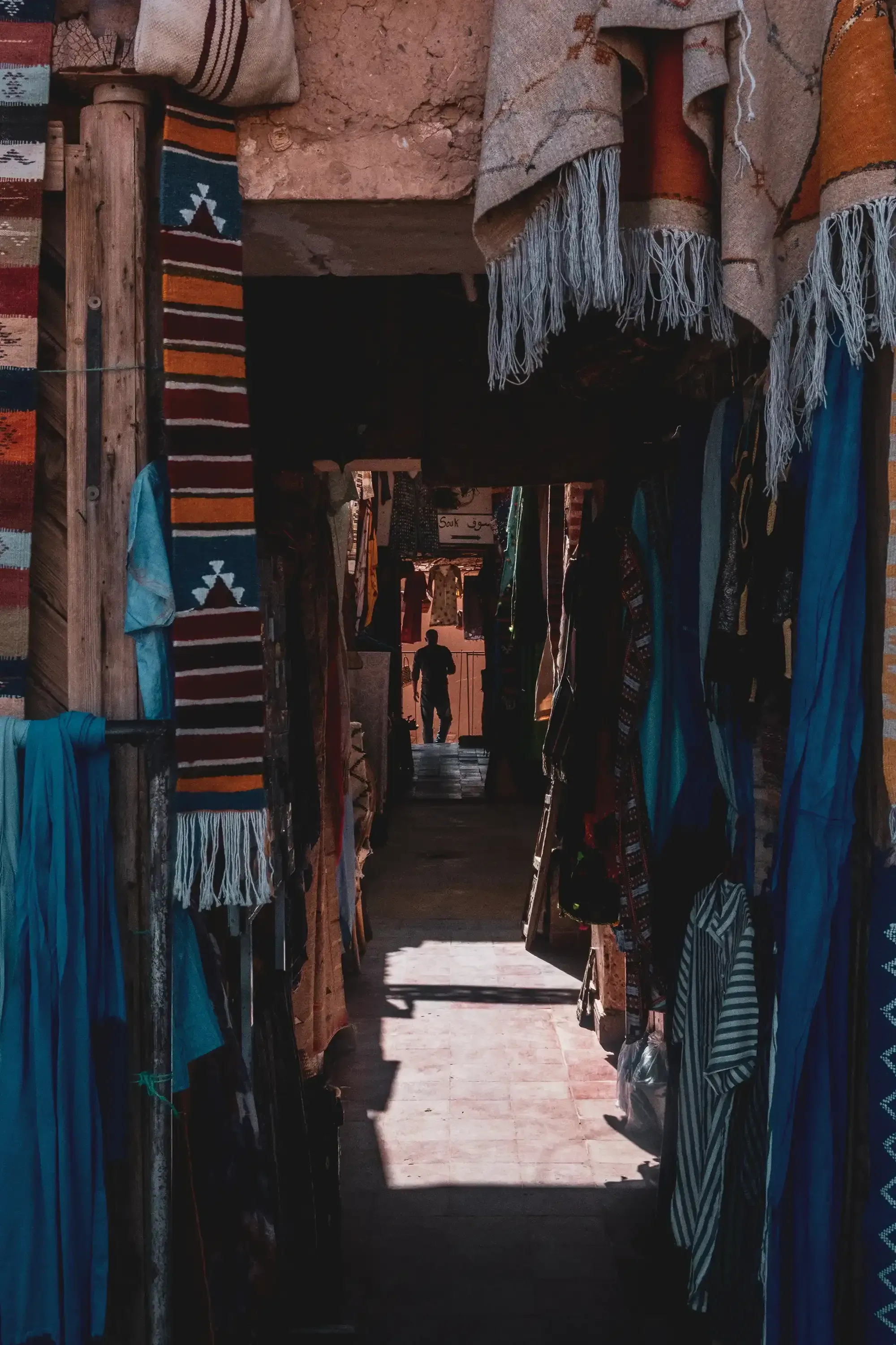 Narrow marketplace street (souk) lined with stalls selling colorful Moroccan textiles.
