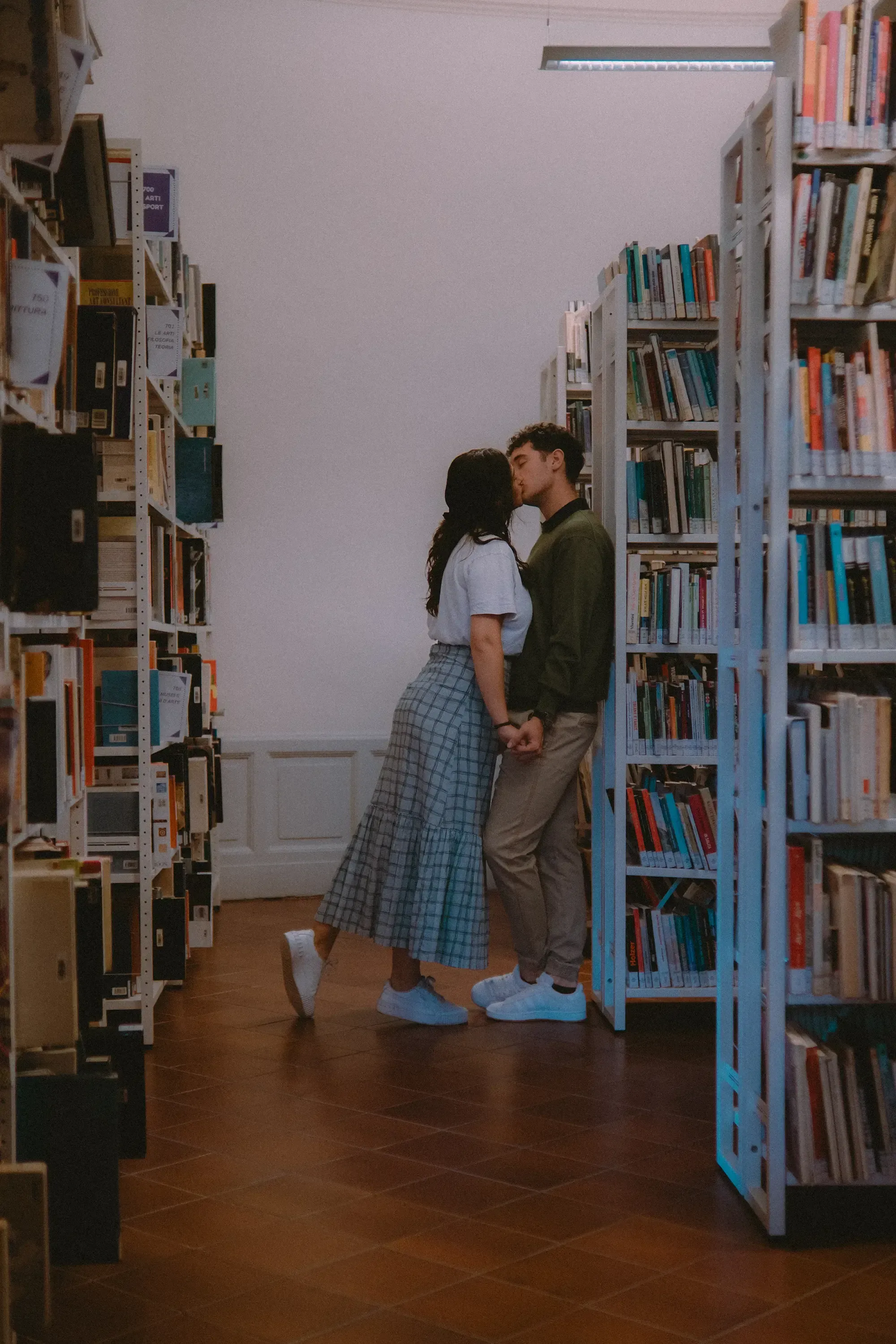 Paulina leaning in to kiss Paolo against a tall wooden library bookshelf.