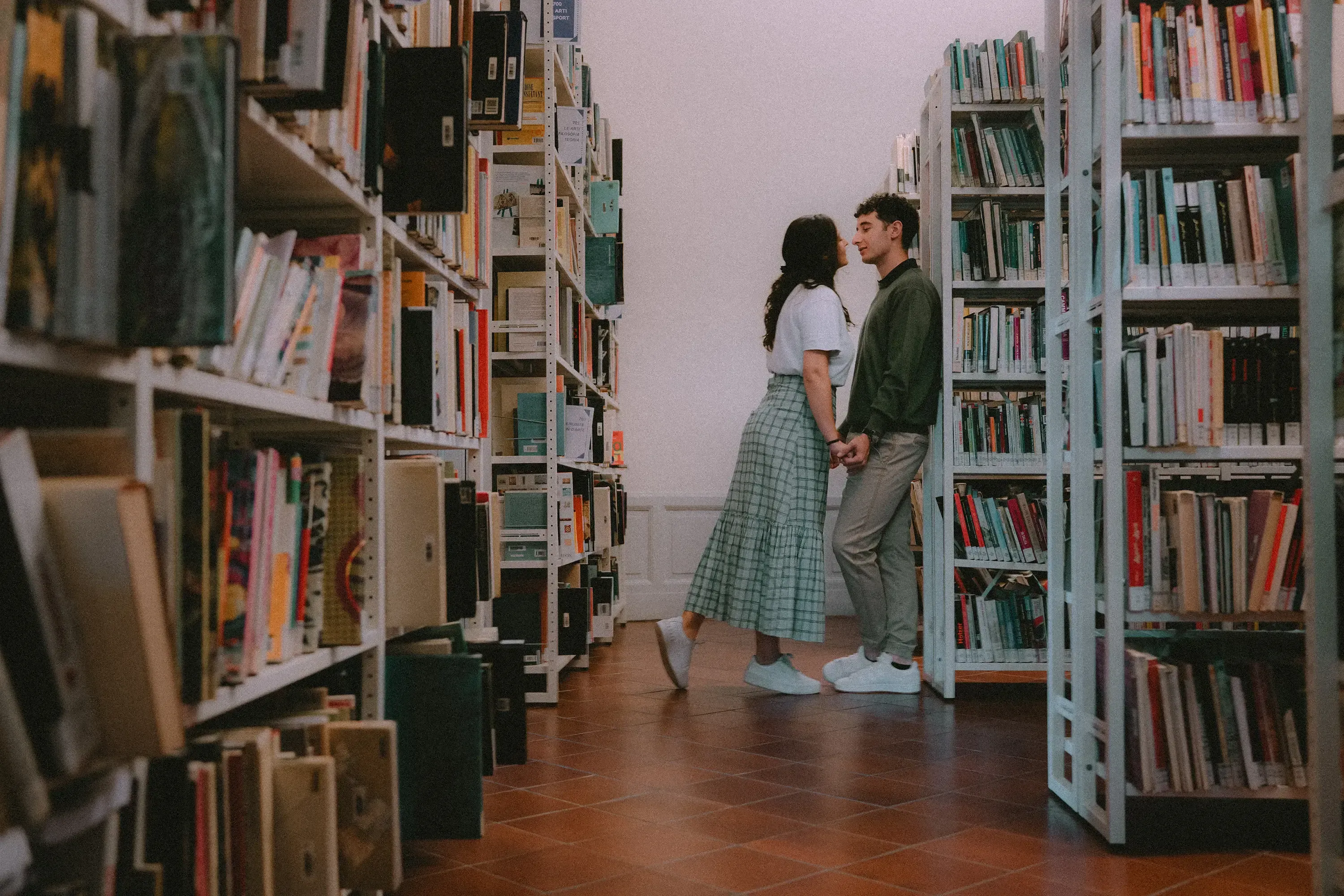 Paolo and Paulina kissing in the background, out of focus, with library books sharp in the foreground.