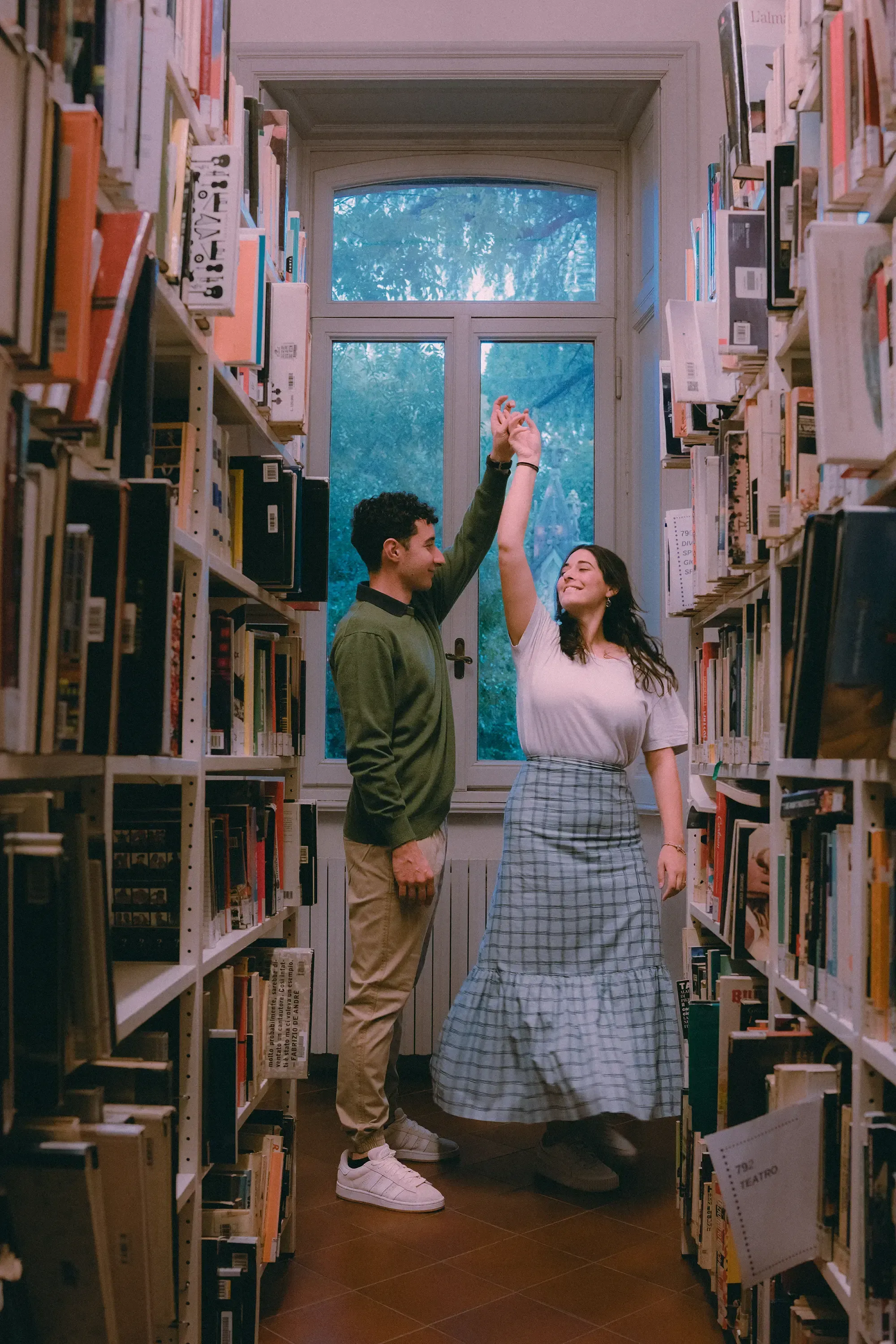 Paolo and Paulina laughing and dancing between the tall rows of library bookshelves.