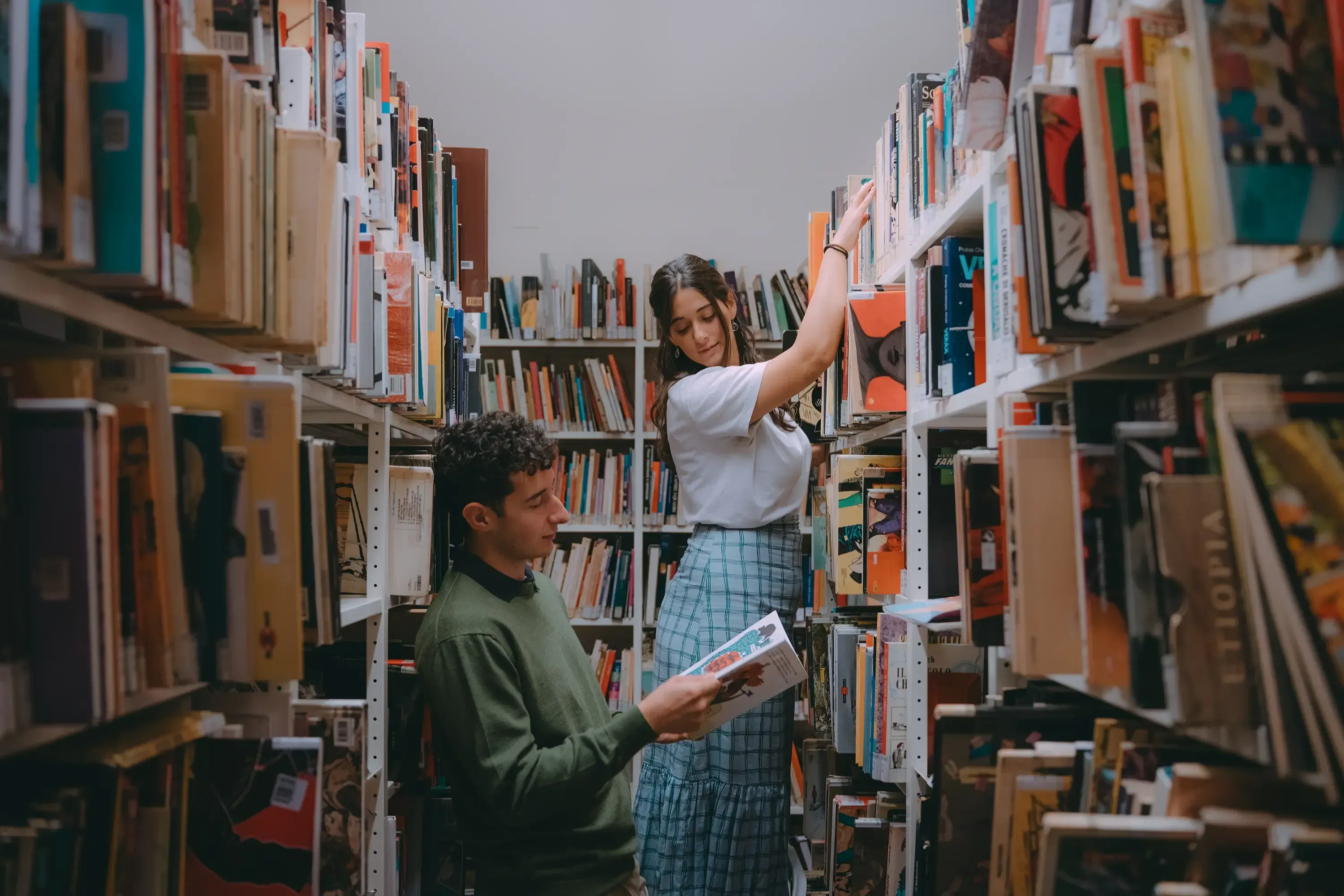 Paulina looking down from a library ladder at Paolo, who is absorbed in reading a book.
