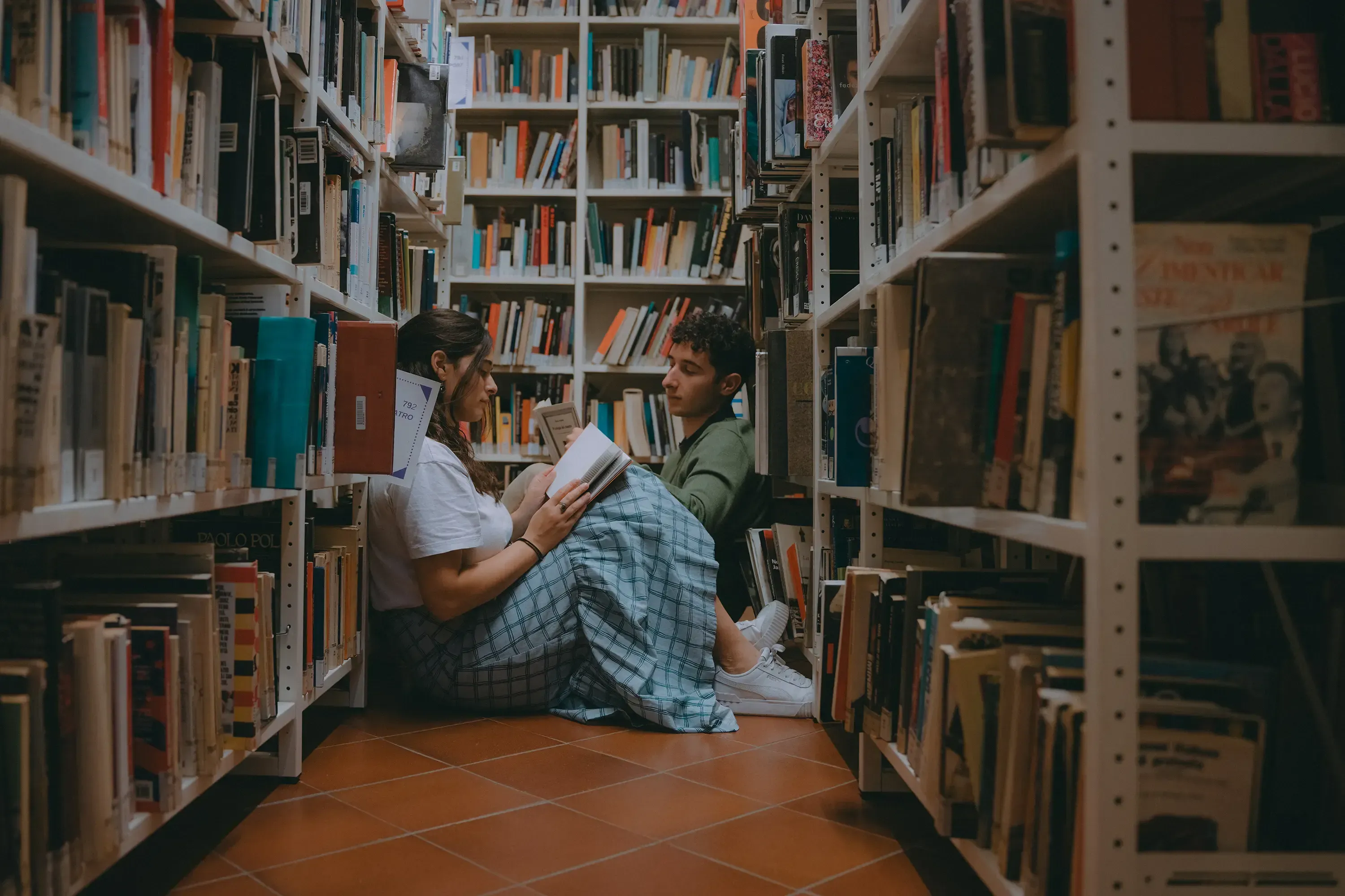 Paolo and Paulina seated together in a library aisle, absorbed in reading.