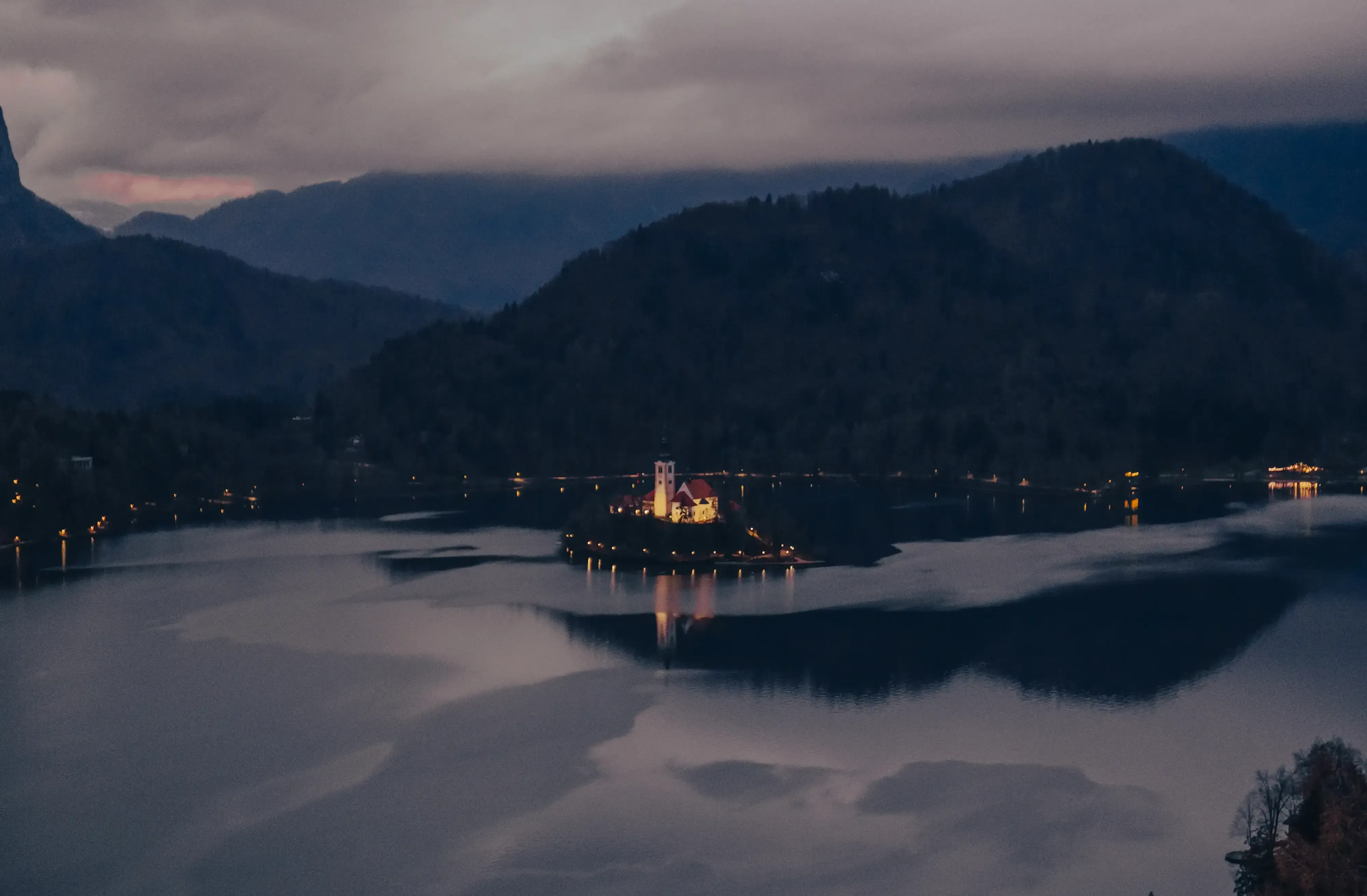 An high-vantage view of the illuminated island church on Lake Bled at night, surrounded by dark water.