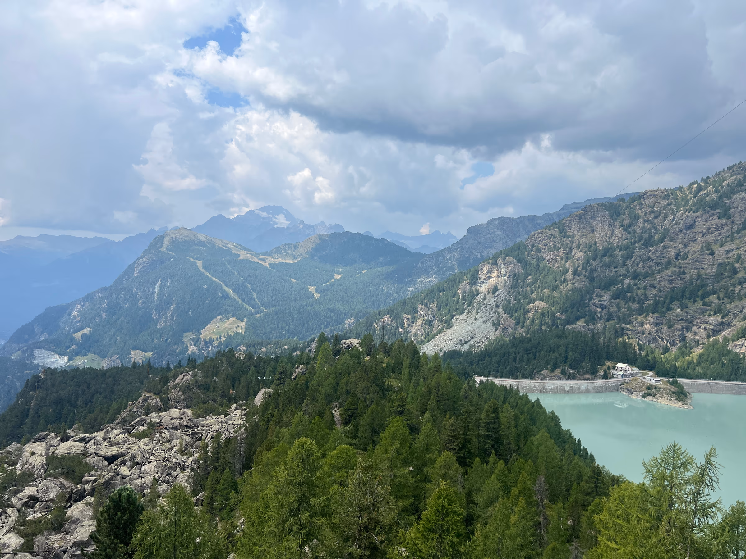 Un bacino montano turchese con la diga, visto da una cresta rocciosa di larici e pini sotto un cielo nuvoloso